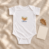 Illustrative lifestyle mockup of one white acorn-name bodysuit. Oliver is an example; card and leaf are props, not included.