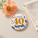 Illustrative celebration tabletop scene of one 2.25 inch birthday pin button. Sarah and 40 are personalized examples; clothing, bags, wrapping and props not included.